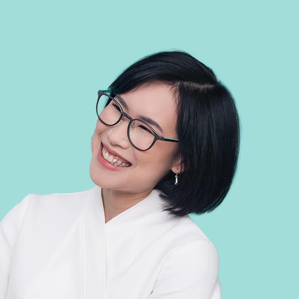 Headshot of Prae Songprasit facing left and smiling widely. She has short shoulder length black hair, and is wearing glasses with a white top. The image sits on a plain Aqua Spray background