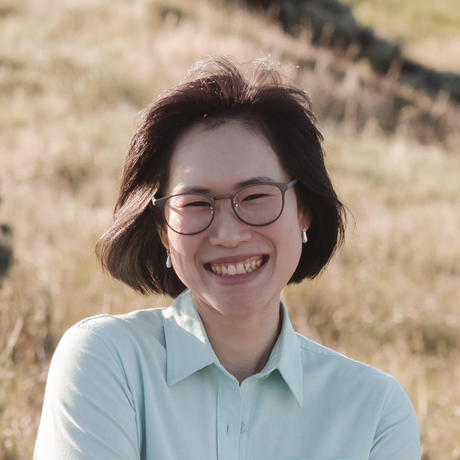 Headshot of Prae smiling widely at the camera. She's wearing a big pair of glasses and a mint green shirt. Her short black hair is fluttering in the wind, and there's a golden plain behind her